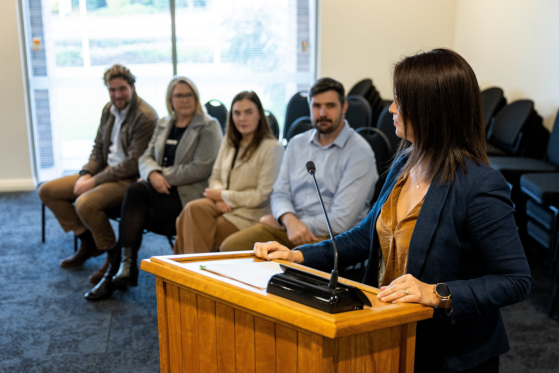 Lady standing at podium presenting with four people watching seated